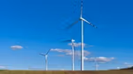A row of wind turbines stand in a ploughed field, set against a bright blue sky