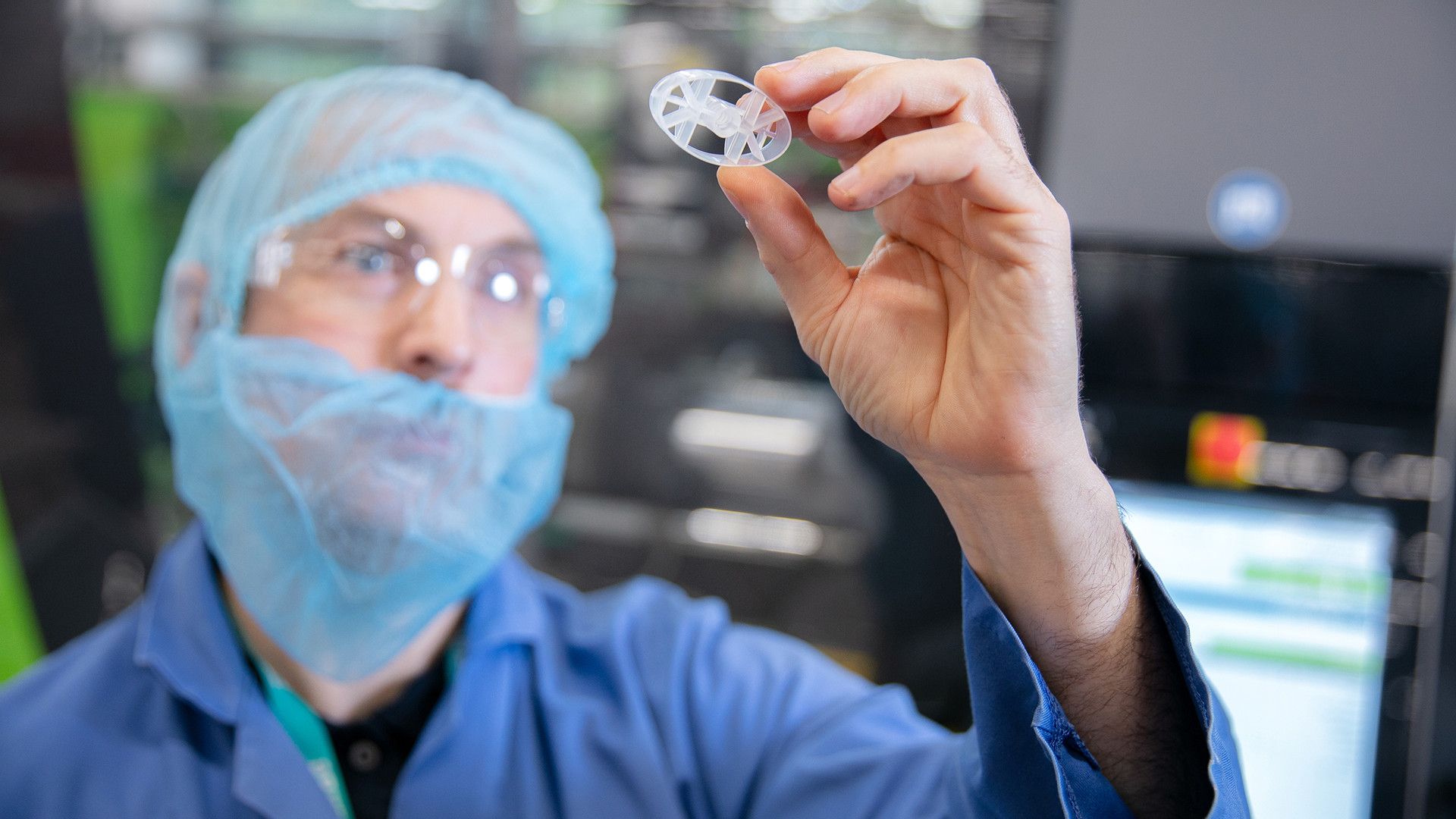 A gloved hand holding a small, circular plastic component in a cleanroom environment with machinery in the background.