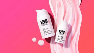 Two white bottles of K18 hair treatment placed on a pink background with swirled cream.