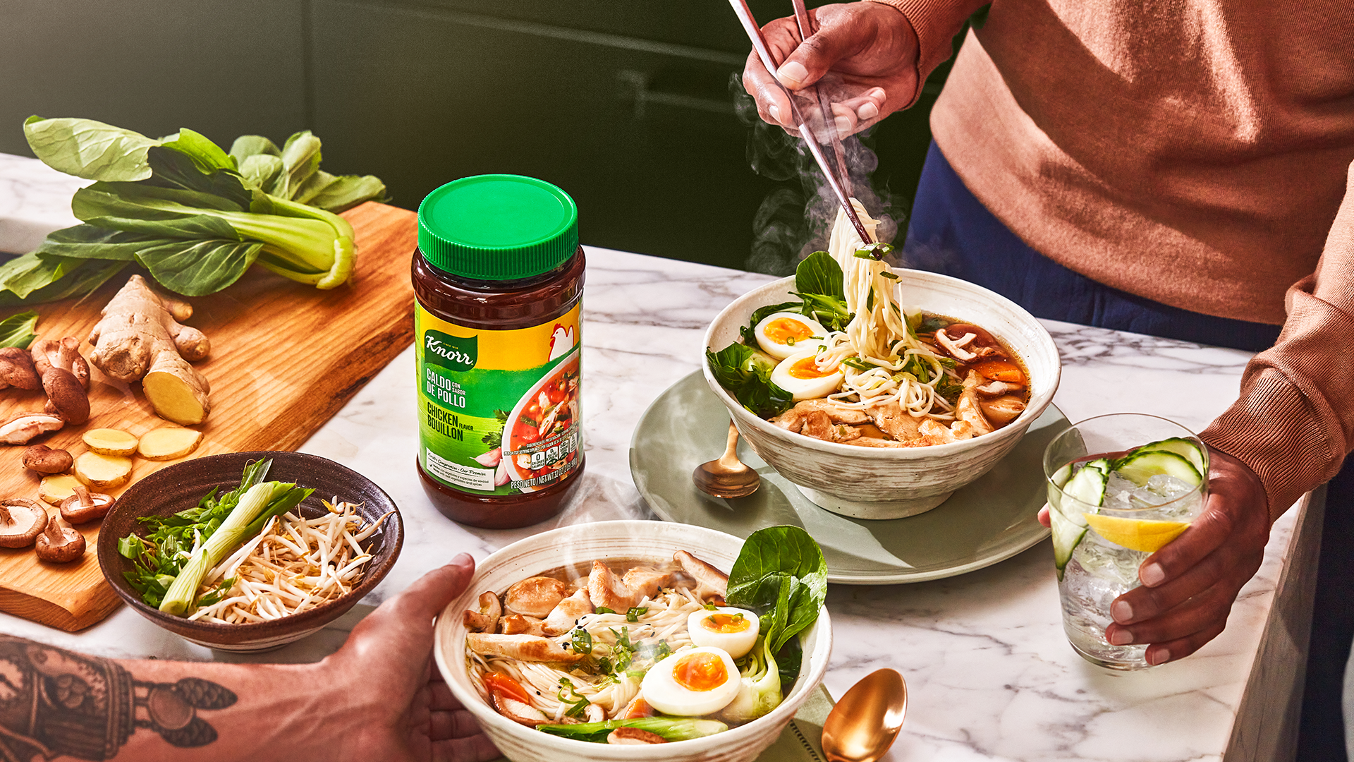 Two people around a table with two full bowls of ramen, some vegetables on a chopping board and a jar of Knorr bouillon.