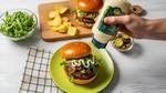 A hand drizzles mayonnaise onto a burger stacked with greens and tomato on a green plate, with chips and pickles on the side.