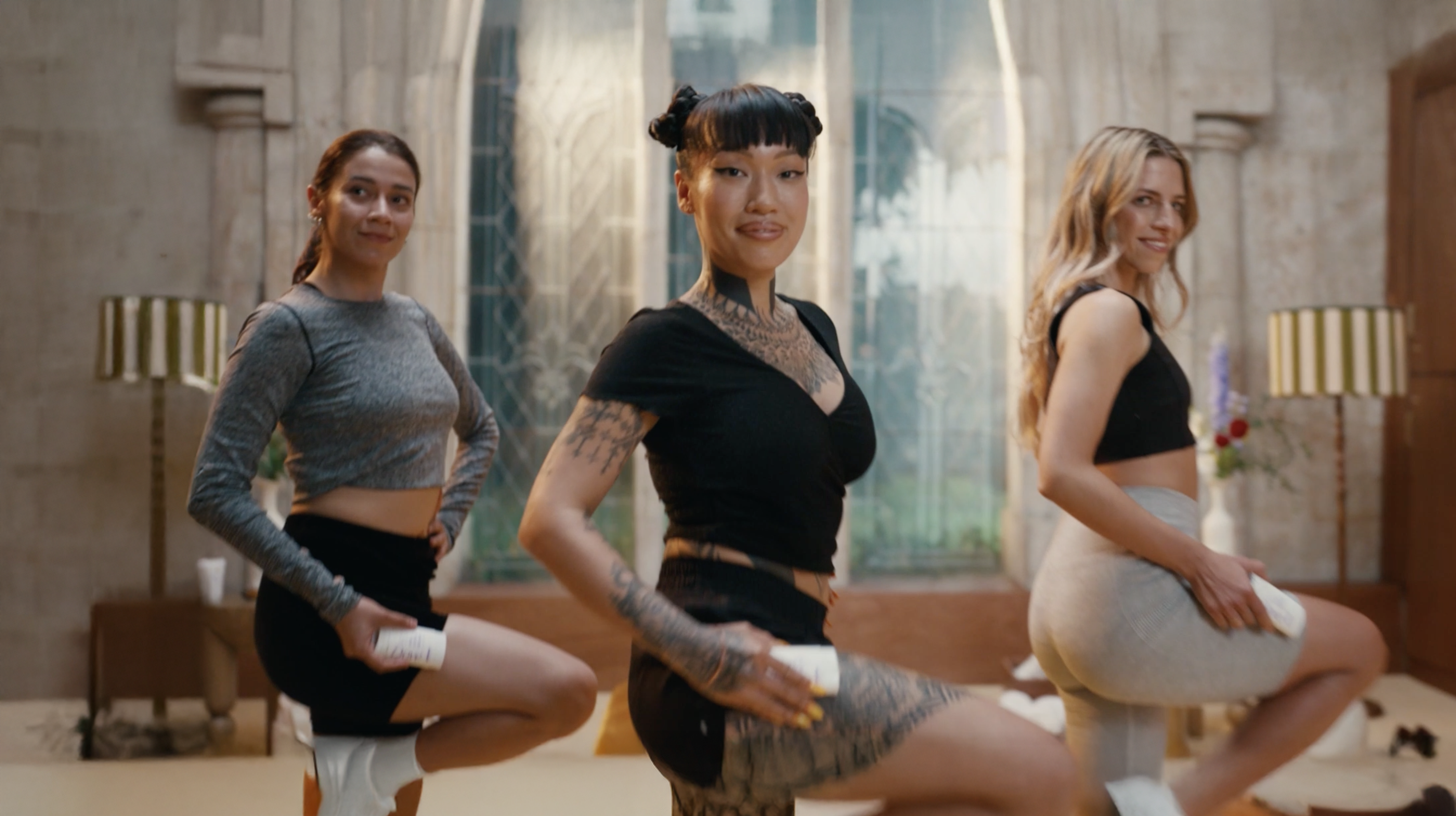 Three individuals performing a fitness or dance routine indoors, each holding a cylindrical object in one hand, with a large window and vintage decor in the background.