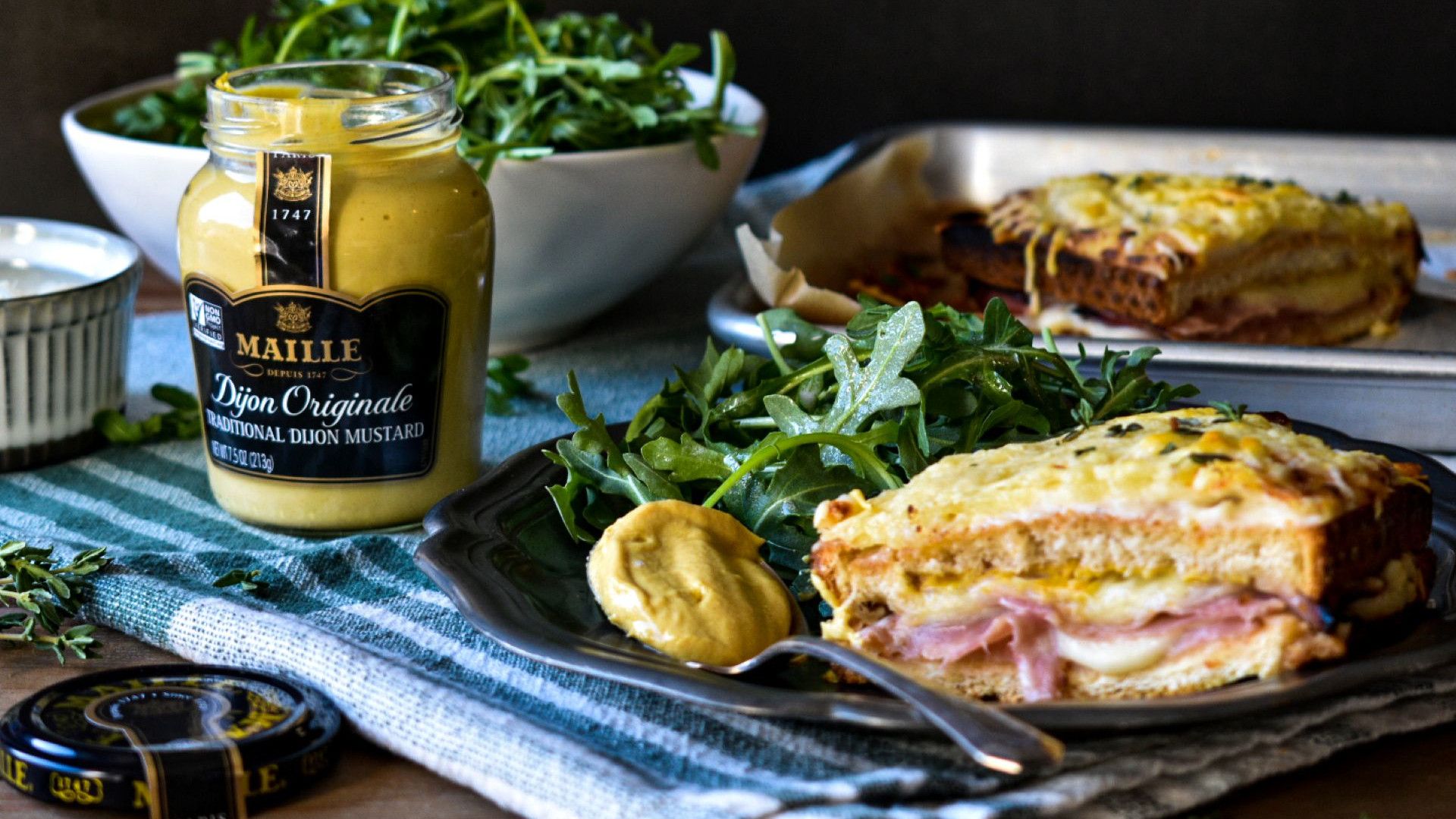 A ham and cheese sandwich served with arugula and Dijon mustard, alongside a jar of Maille mustard.